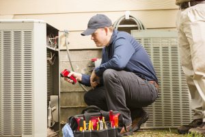 Mid-adult repairman works on a home's air conditioner unit outdoors. Man center is working on the unit using tools in his toolbox. Other man to right. They both wear uniforms.